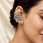 A close-up of a woman's ear wearing a decorative, multi-colored ear cuff with gold-toned metal and green, pink, and blue stones, set against a soft beige satin background.