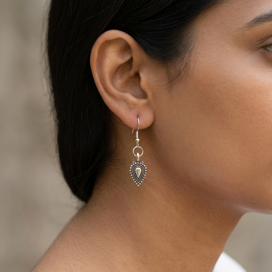 A close-up of a woman's ear wearing a silver dangle earring with a heart-shaped charm featuring a central oval inset and beaded border details.