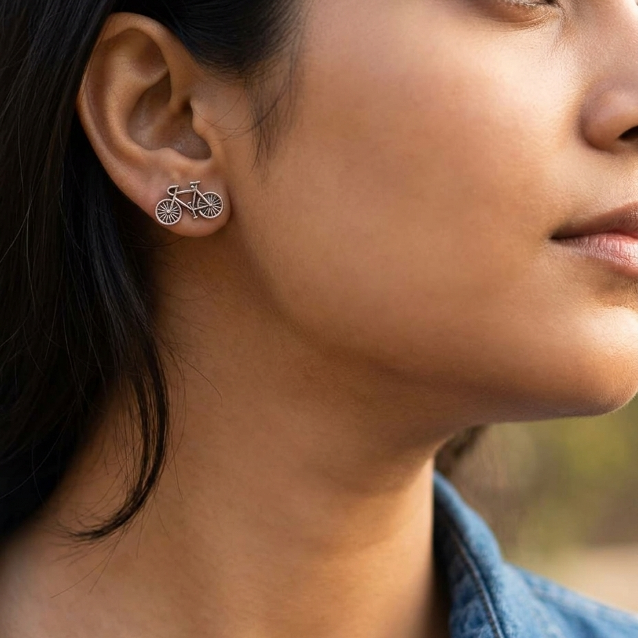 A close-up of a woman's ear wearing a small silver bicycle stud earring, with her face, neck, and denim shirt visible against a softly blurred outdoor background.