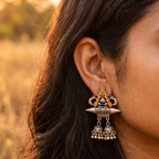 A close-up of a woman's ear wearing an ornate traditional Indian jhumka earring in silver-toned metal with yellow-orange stone accents, set against a softly blurred warm outdoor background.