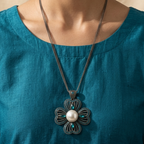 A close-up of a woman wearing a dark silver flower-shaped pendant necklace with a white pearl center and small turquoise accents, paired with a teal top against a warm beige background.