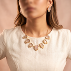 A close-up of a woman wearing a gold-tone statement necklace with rectangular pale pink stones and matching drop earrings, paired with a cream linen top against a soft beige background.