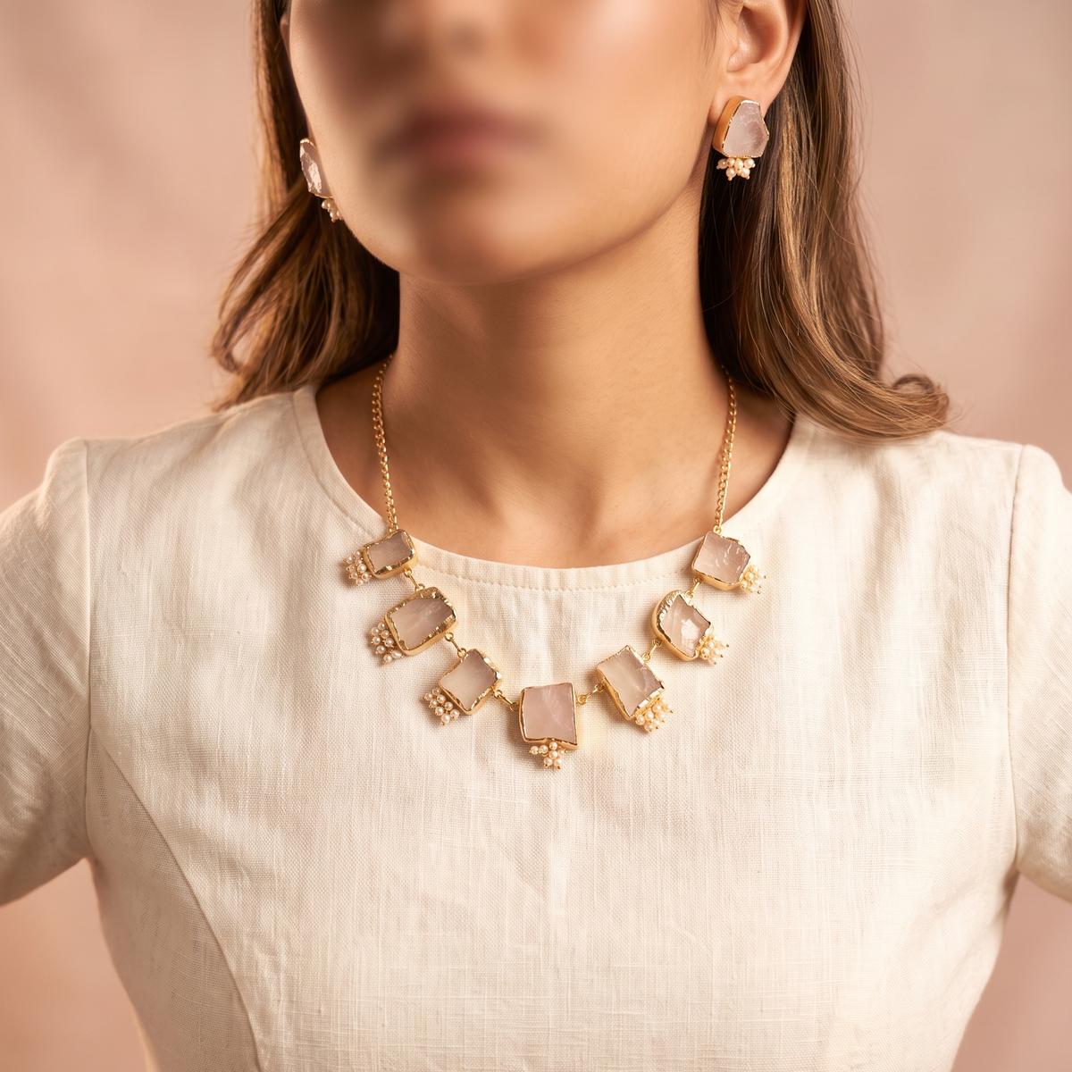 A close-up of a woman wearing a gold-tone statement necklace with rectangular pale pink stones and matching drop earrings, paired with a cream linen top against a soft beige background.