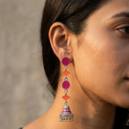 A close-up of a woman wearing a long, multi-tiered dangle earring. The earring features magenta-pink circular and diamond-shaped enamel details with a silver-toned bell-shaped charm at the bottom.