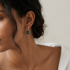 A close-up of a woman wearing a small gold hoop earring with a round green drop pendant, paired with a white top against a soft light background.