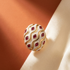 A close-up of an oval ring with a white enamel face, gold wavy metal borders, and red square gemstones, resting on draped cream satin fabric against a warm brown background.