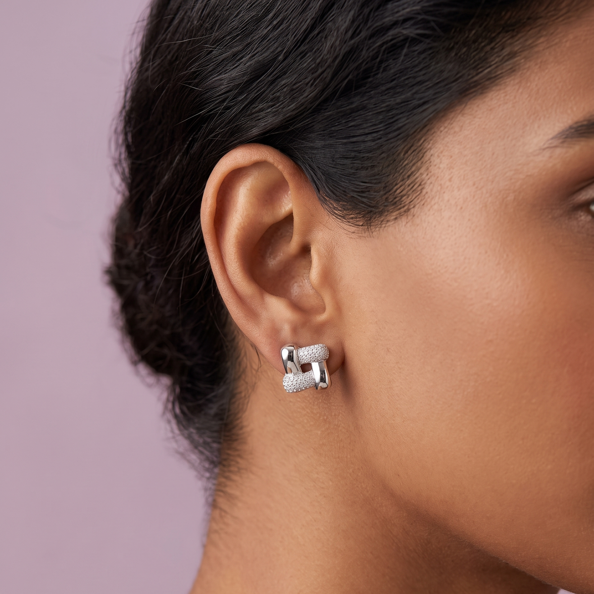 A close-up side profile of a model wearing a pair of silver huggie hoop earrings with textured, diamond-like pavé detailing, set against a solid dusty lavender background.