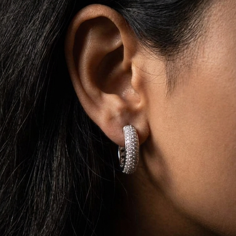 A close-up side profile of a person's ear wearing a small silver hoop earring set with clear stones, against dark hair and warm skin tones.