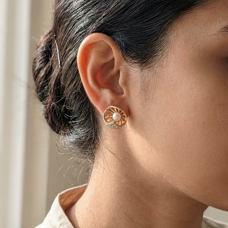 A close-up side profile of a person wearing a small gold-toned dangle earring with a white pearl at the center, styled with dark hair tied back and a beige top.