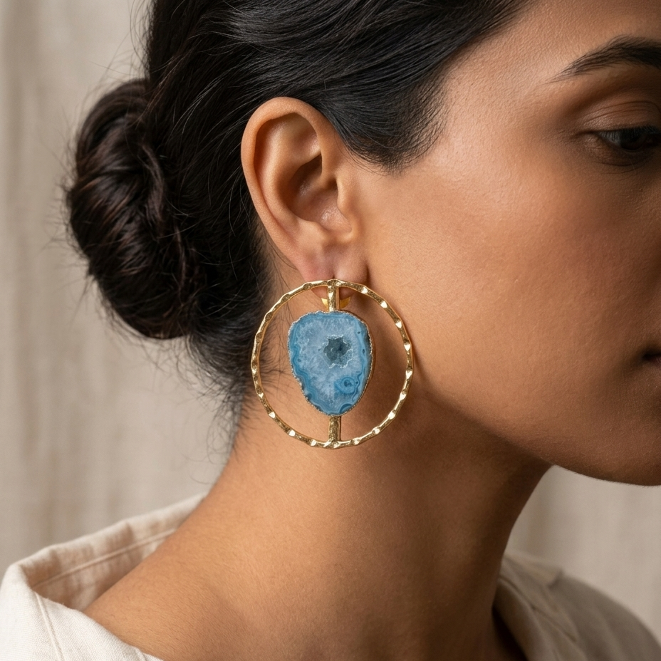 A close-up side profile of a woman wearing a large gold hoop earring with a blue agate slice pendant, paired with a light beige linen shirt.