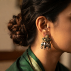 A close-up side profile of a woman wearing a silver-toned elephant earring adorned with green and gold decorative details, paired with a dark green garment.