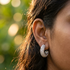 A close-up side profile of a woman wearing a silver crescent-shaped hoop earring paved with clear stones, set against a soft, blurred green bokeh background.