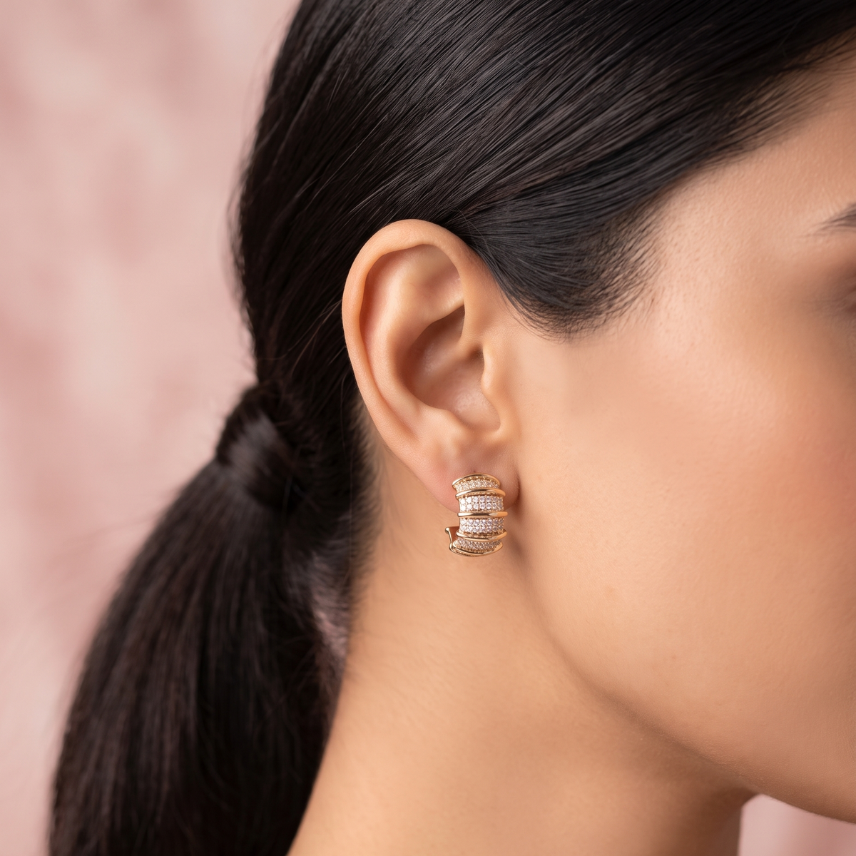 A close-up side profile of a woman wearing a small curved hoop earring with multiple rows of clear stones set in a warm gold-tone metal, against a soft pink background.
