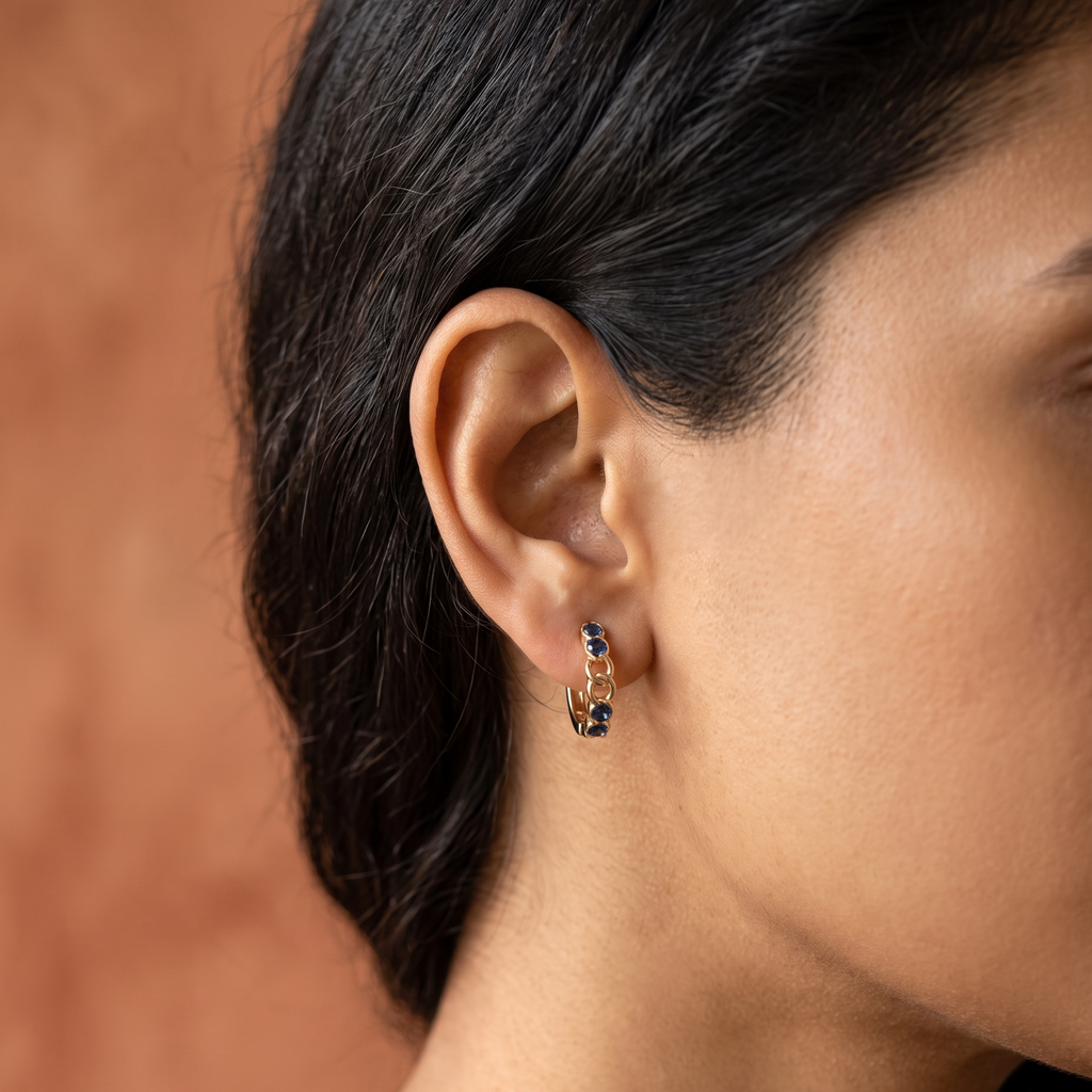 A close-up side profile of a woman wearing a small gold huggie hoop earring adorned with dark blue gemstones, set against a warm, textured terracotta background.