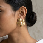 A close-up side profile of a woman wearing a textured gold flower-shaped earring with a small white pearl at the center, paired with a white collared shirt against a neutral beige background.