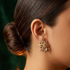 A close-up side profile of a woman with dark hair in a bun wearing a gold-toned, flower-like stud earring with sparkling clear stones against a dark green background.
