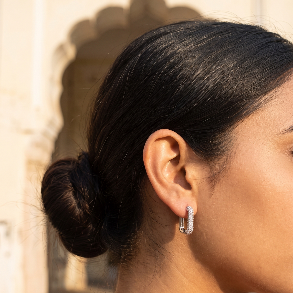 A close-up side profile of a woman with her hair in a bun wearing a silver huggie hoop earring with pavé-set clear stones, set against a warm, sunlit architectural background.