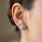 A close-up side view of a woman's ear wearing a round silver-toned stud earring with a textured, diamond-like pavé setting.