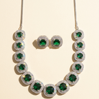 A gold-tone necklace and matching stud earrings set with emerald-cut green gemstones surrounded by clear pavé stones, arranged on a light beige background.