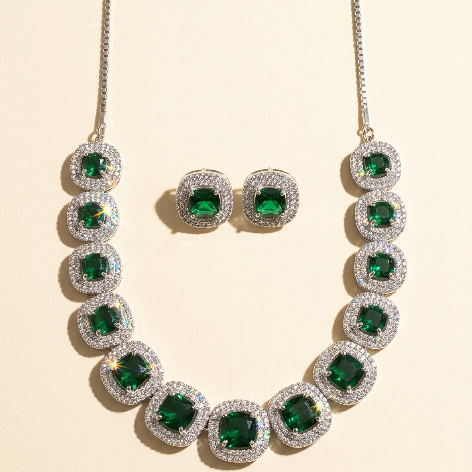 A gold-tone necklace and matching stud earrings set with emerald-cut green gemstones surrounded by clear pavé stones, arranged on a light beige background.