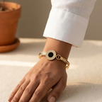 A gold-toned bangle bracelet with a central black oval stone and a textured gold band, shown worn on a person's wrist with a white shirt cuff visible.