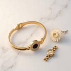 A gold-toned bangle bracelet with a central oval black inlay surrounded by small clear stones, displayed on a white marble surface alongside a small gold watch and a decorative white flower.