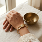 A gold-toned bangle bracelet with colorful enamel details is worn on a person's wrist, shown in a styled lifestyle setting with a small brass bowl on a linen tablecloth.