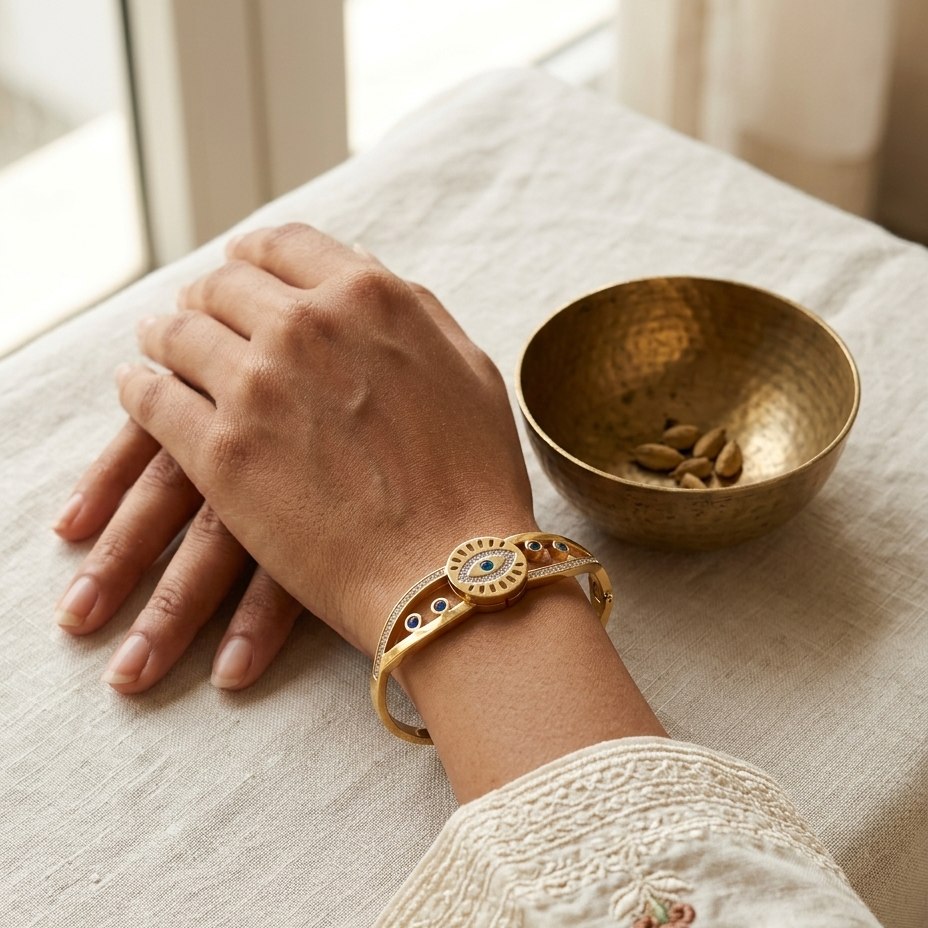 A gold-toned bangle bracelet with colorful enamel details is worn on a person's wrist, shown in a styled lifestyle setting with a small brass bowl on a linen tablecloth.