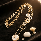 A gold-toned chain necklace with white circular shell-like disc accents and a safety-pin style connector, displayed on a black velvet surface with small dried flowers and a white stone prop.