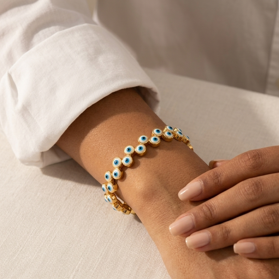 A gold-toned charm bracelet with blue and white evil eye beads worn on a person's wrist, paired with a white long-sleeve shirt.