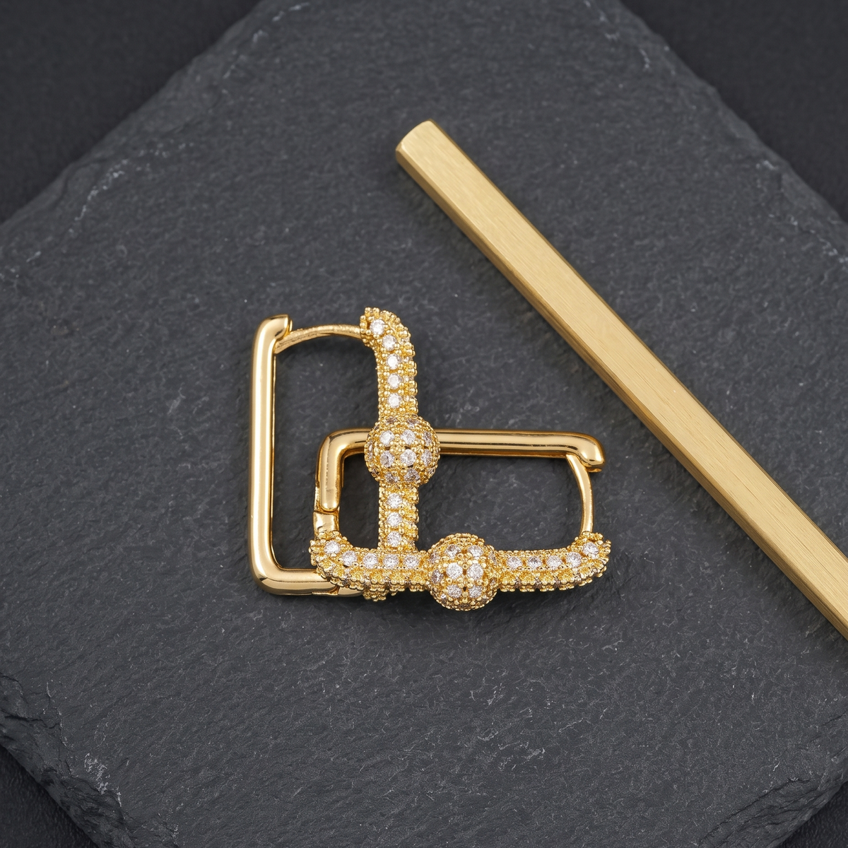 A gold-toned earring with pavé-set clear stones, displayed on a dark slate surface next to a gold bar.