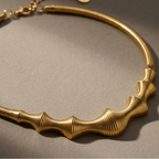 A gold-toned metal collar necklace with a ribbed, wavy central design and a textured snake-style band, photographed on a dark gray surface with dramatic lighting.