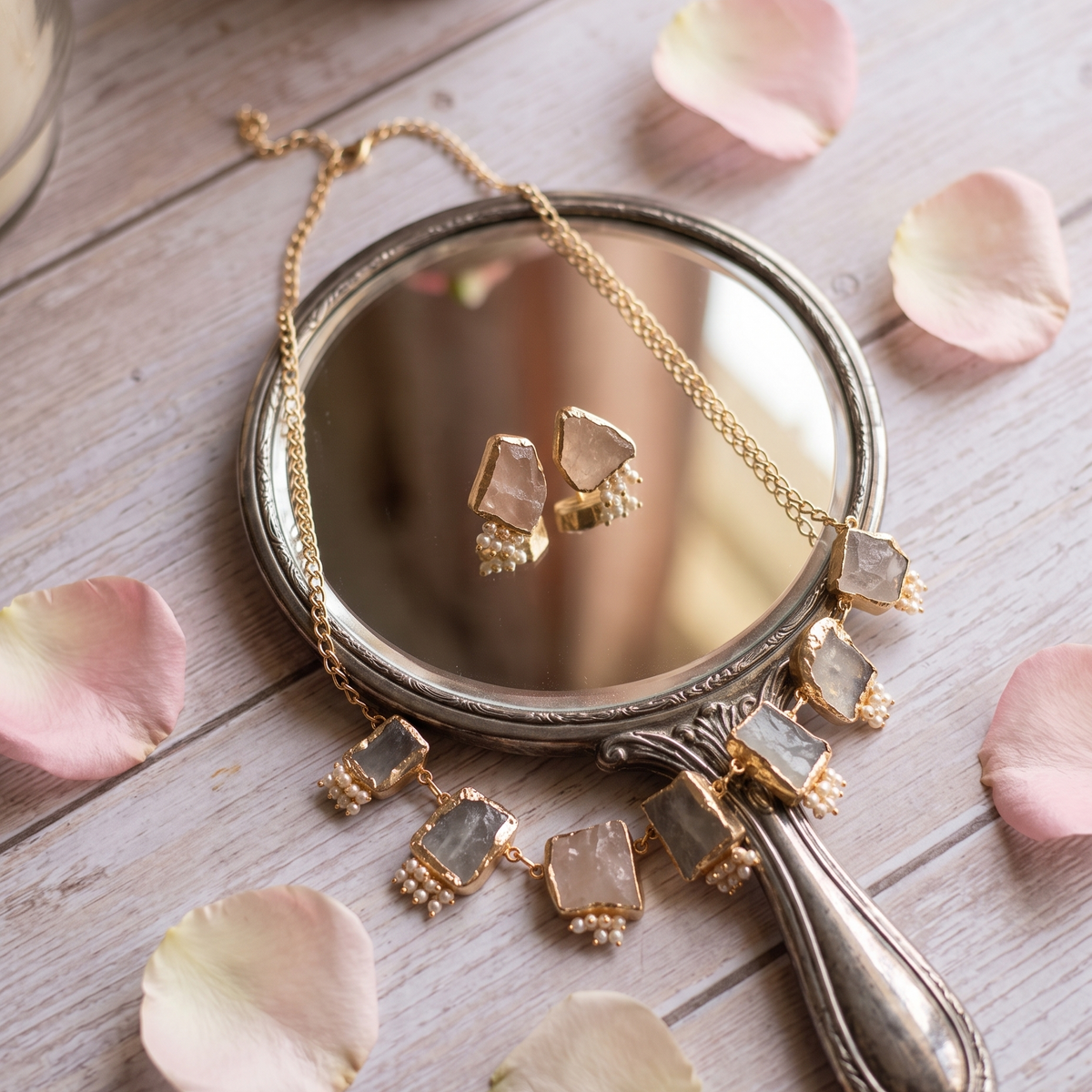 A gold-toned necklace with rectangular clear gemstone charms draped over a vintage-style silver mirror, styled on a light wood surface with scattered pink rose petals.