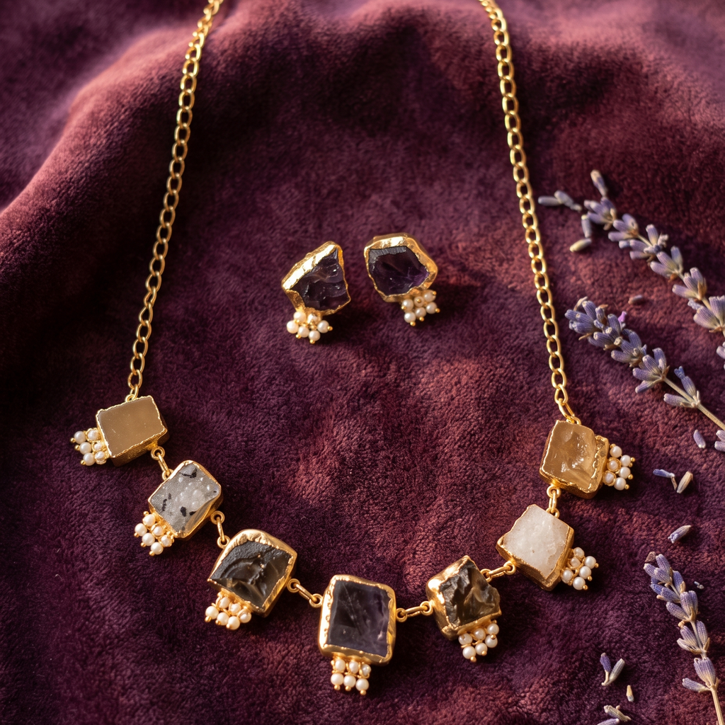 A gold-toned statement necklace and matching stud earrings displayed on a deep burgundy velvet fabric, accented with dried lavender sprigs. The necklace features multiple rectangular gemstone charms in black, white, and smoky tones, each decorated with small white bead clusters.