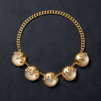 A gold chain necklace featuring five large, round cream-colored beads with dripping gold accents, displayed on a dark textured background.