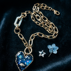 A gold chain necklace with a heart-shaped blue pendant featuring an ocean-themed pattern, displayed on a dark velvet background alongside a blue flower pin and a star-shaped charm.