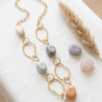 A gold chain necklace with irregular stone beads in cream, gray, mint green, peach, and lavender, styled on a white textured fabric next to a dried pampas grass stem and three loose stones.