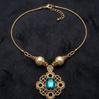 A gold chain necklace with two cream pearl beads, a small clear crystal accent, and a central gold filigree pendant featuring a rectangular teal-blue gemstone, displayed on a dark textured background.