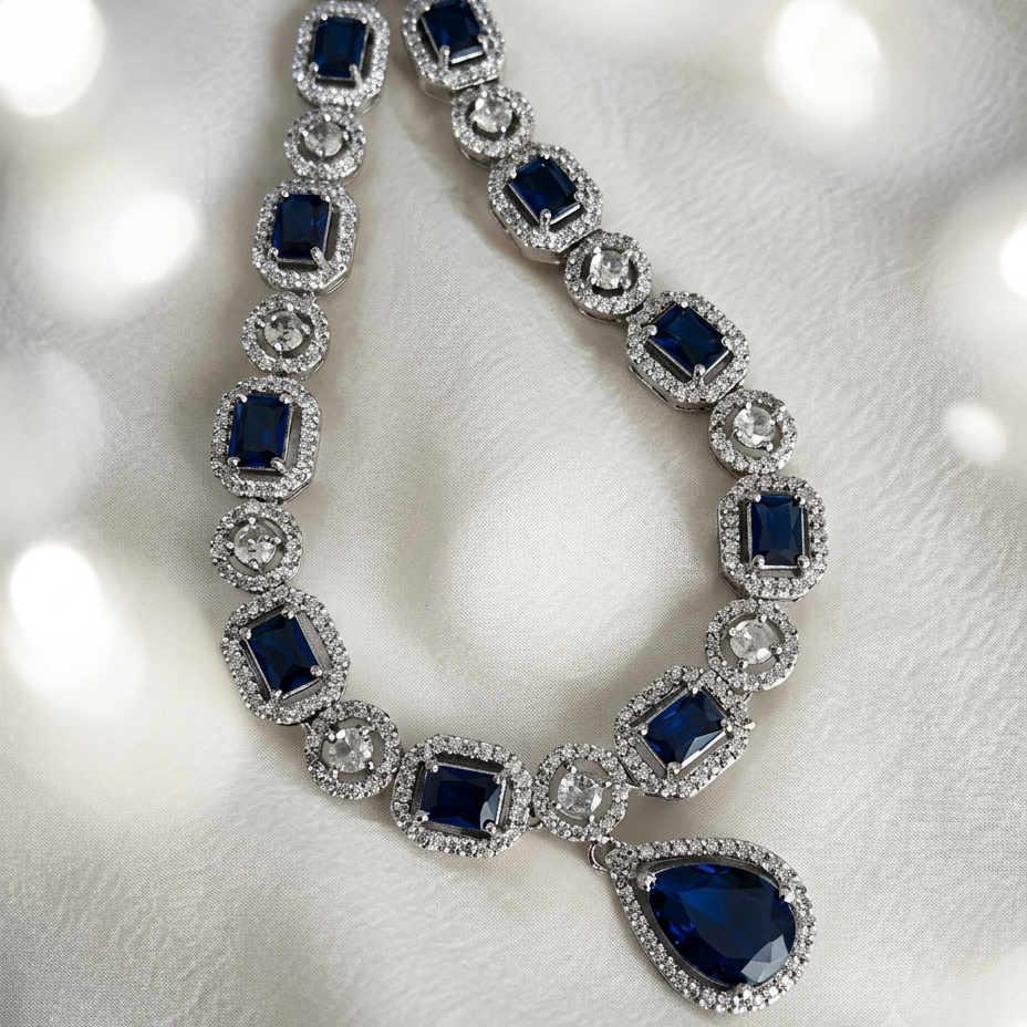 A luxury silver-toned necklace featuring deep blue rectangular and teardrop-cut gemstones, each surrounded by a halo of small clear stones, displayed on a soft white textured fabric background with soft bokeh light effects.