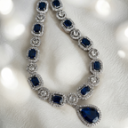 A luxury silver-toned necklace featuring deep blue rectangular and teardrop-cut gemstones, each surrounded by a halo of small clear stones, displayed on a soft white textured fabric background with soft bokeh light effects.