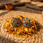A pair of bohemian-style geometric earrings with teal, orange, and bronze details, displayed on a bed of dried flowers over a woven mat.