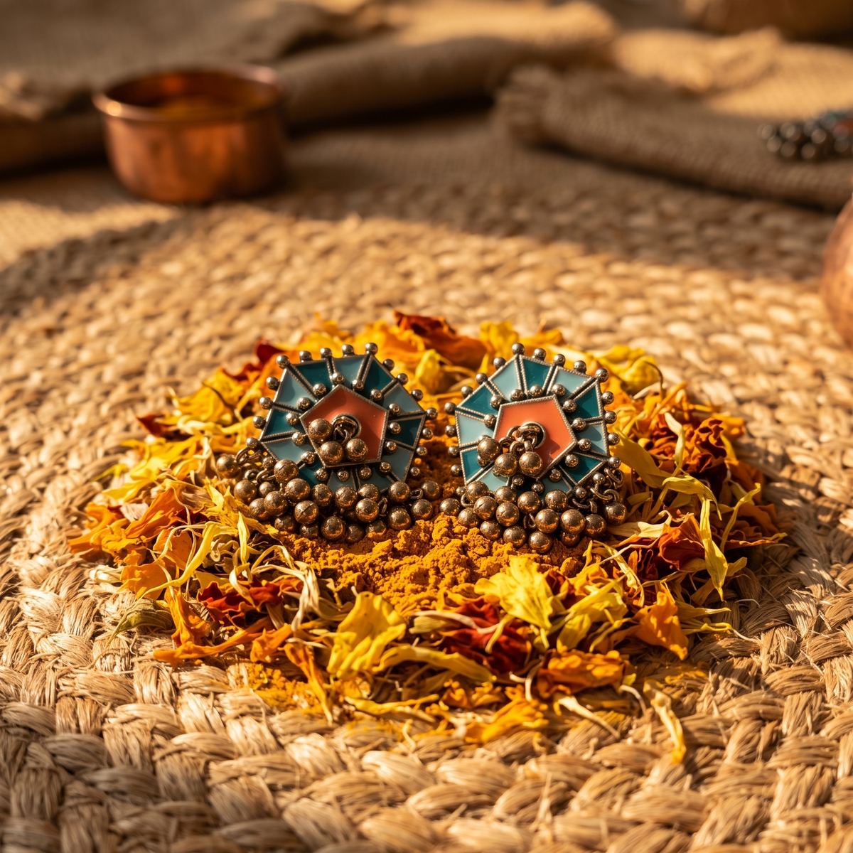 A pair of bohemian-style geometric earrings with teal, orange, and bronze details, displayed on a bed of dried flowers over a woven mat.