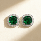 A pair of cushion-cut emerald green gemstone stud earrings with diamond halo settings, displayed on a textured beige surface with soft diagonal shadows.