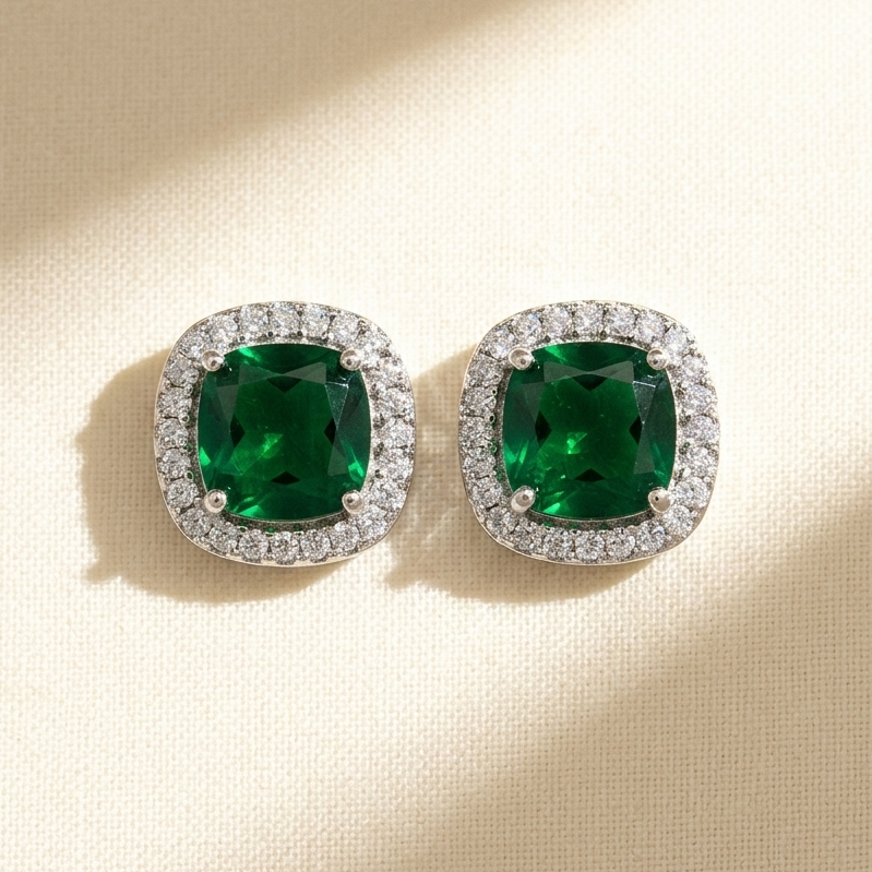 A pair of cushion-cut emerald green gemstone stud earrings with diamond halo settings, displayed on a textured beige surface with soft diagonal shadows.