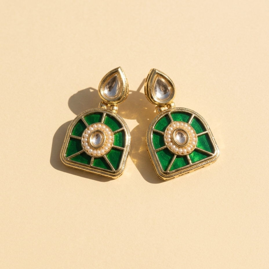 A pair of gold-plated kundan earrings with vibrant green enamel inlay, a central white pearl, and a teardrop-shaped clear stone at the top, displayed on a warm beige background.