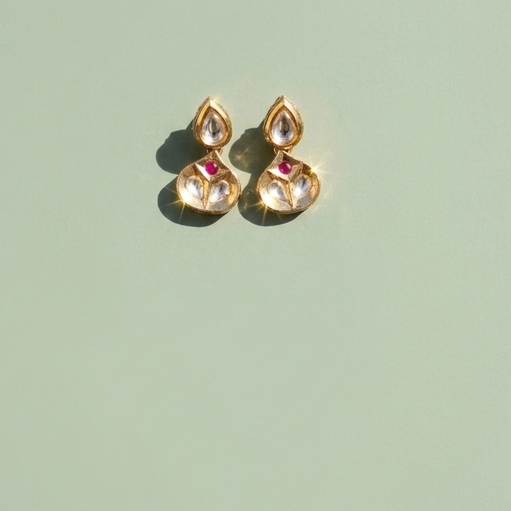 A pair of gold-toned Indian jhumka earrings with clear teardrop crystals and small pink gemstone accents, displayed on a solid sage green background.