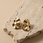 A pair of gold-toned, abstractly textured stud earrings displayed on a rough, light beige stone slab against a solid beige background.
