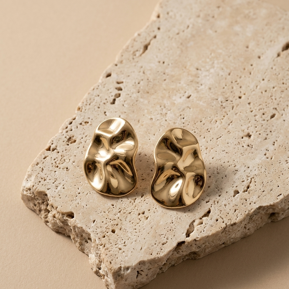 A pair of gold-toned, abstractly textured stud earrings displayed on a rough, light beige stone slab against a solid beige background.