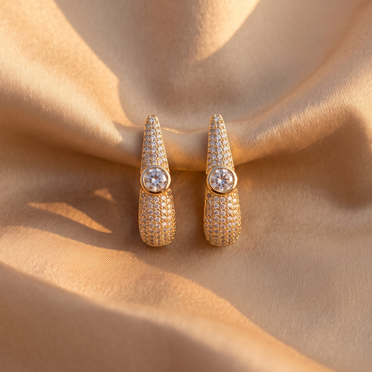 A pair of gold-toned drop earrings with pavé-set clear stones and a central round clear gemstone, displayed on draped champagne satin fabric.