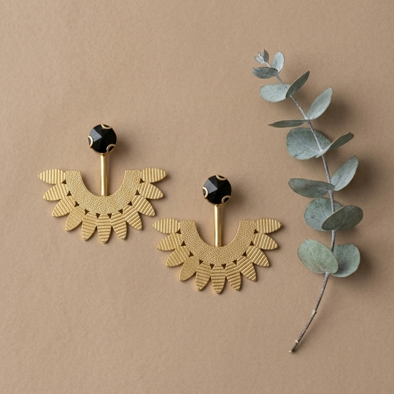 A pair of gold-toned statement earrings with a scalloped fan design and a small black bead at the top, displayed on a warm beige background next to a sprig of eucalyptus.